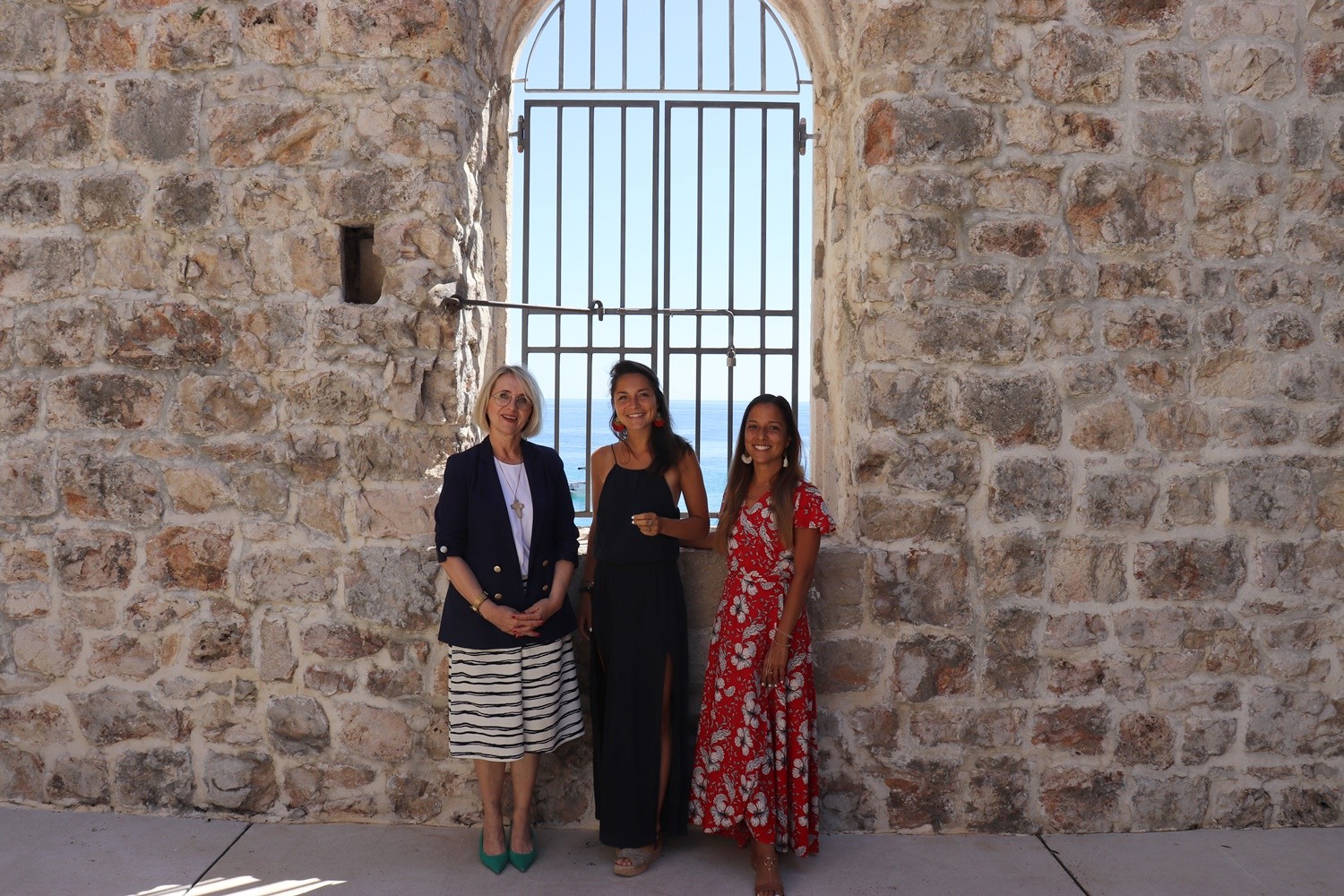 Three women standing in front of a barred window with a sea view in a stone building