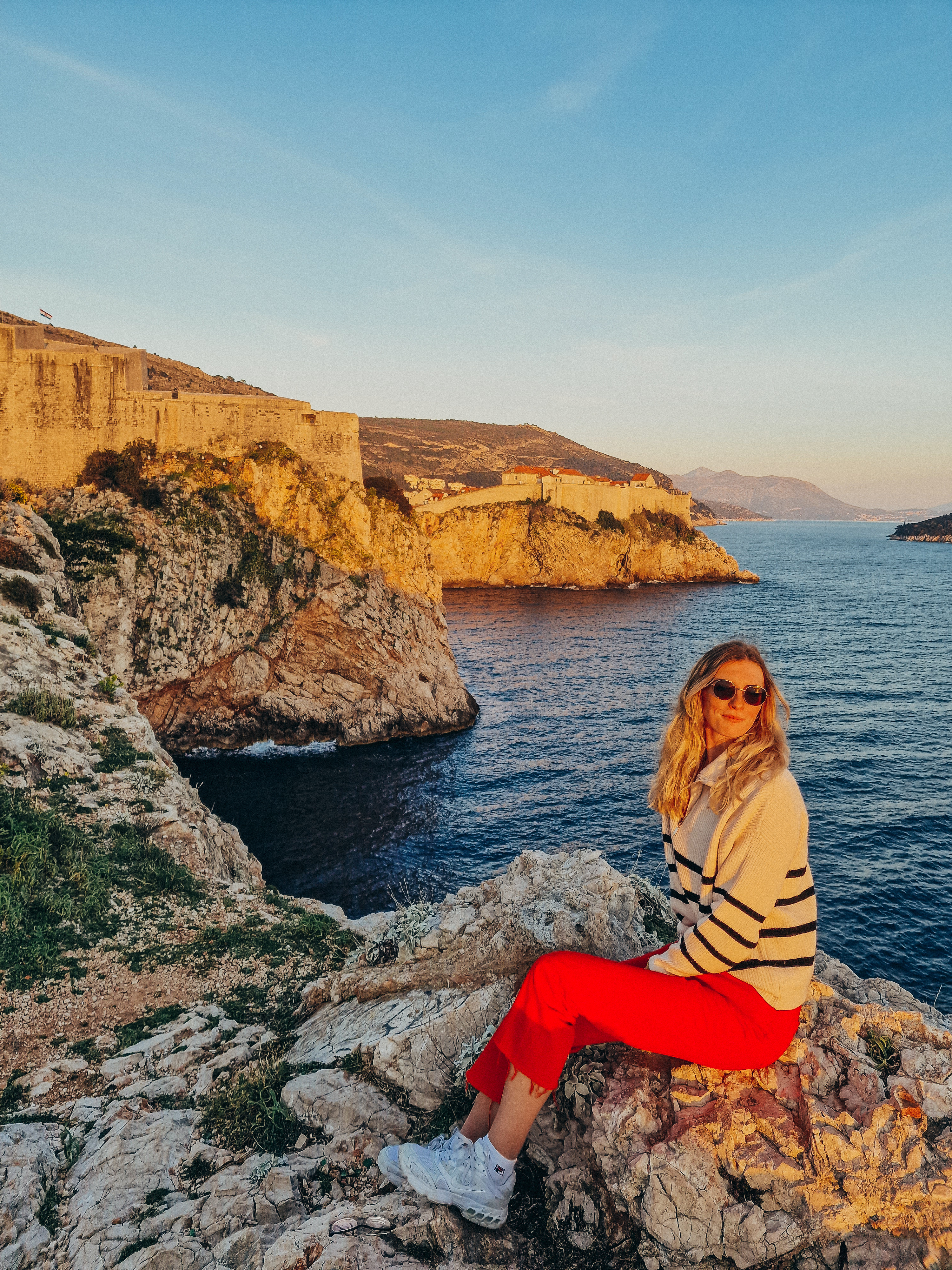 Helena Bradbury sitting on seaside rocks at sunset, with Dubrovnik's fortress walls across the water