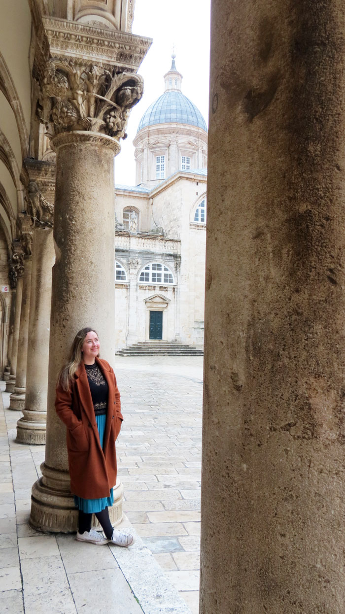 Han Talbot standing under a stone colonnade in Dubrovnik Old Town, with a domed church behind her