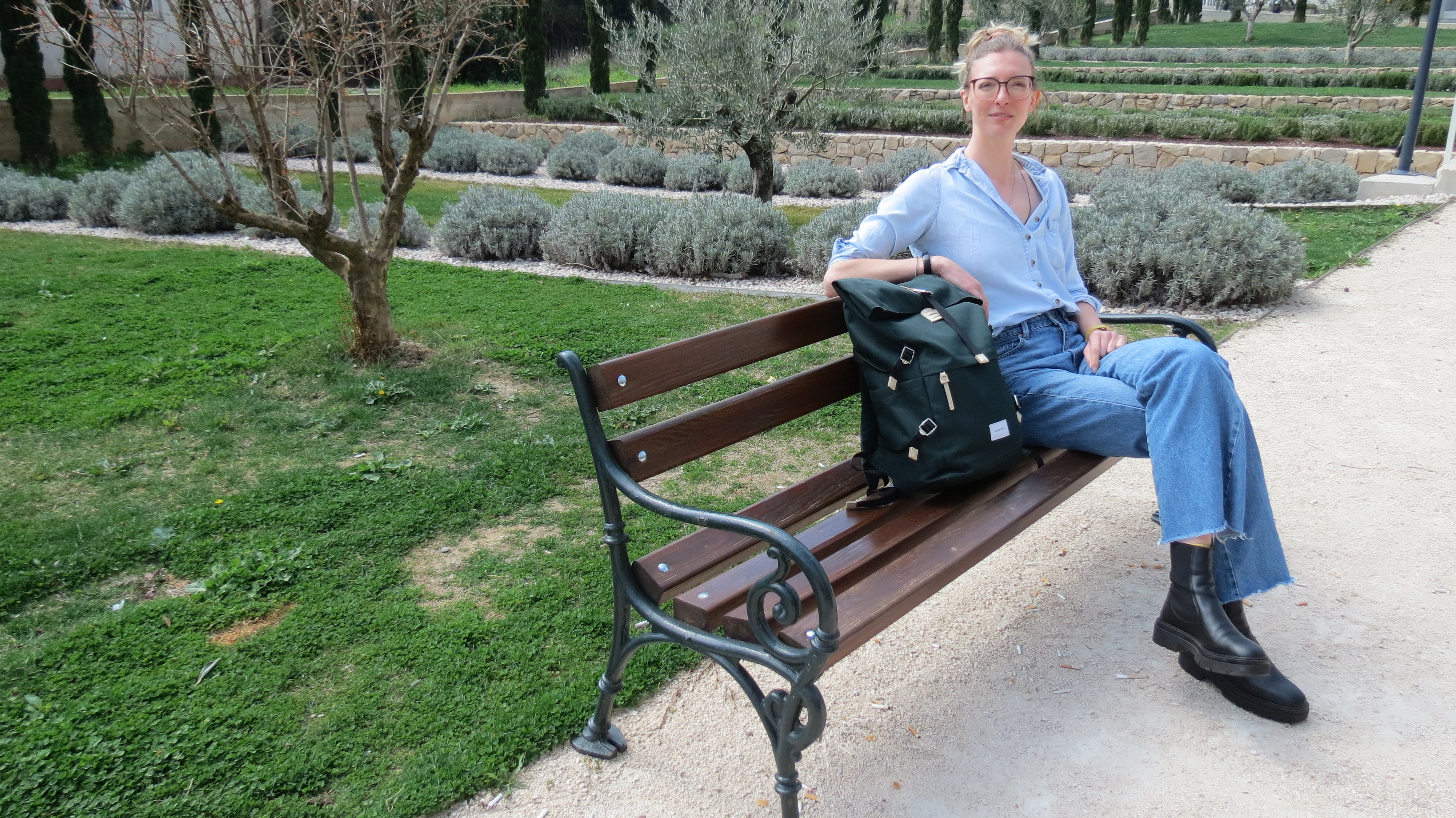 Helena Bradbury sitting on a park bench with a green backpack, beside olive trees and lavender