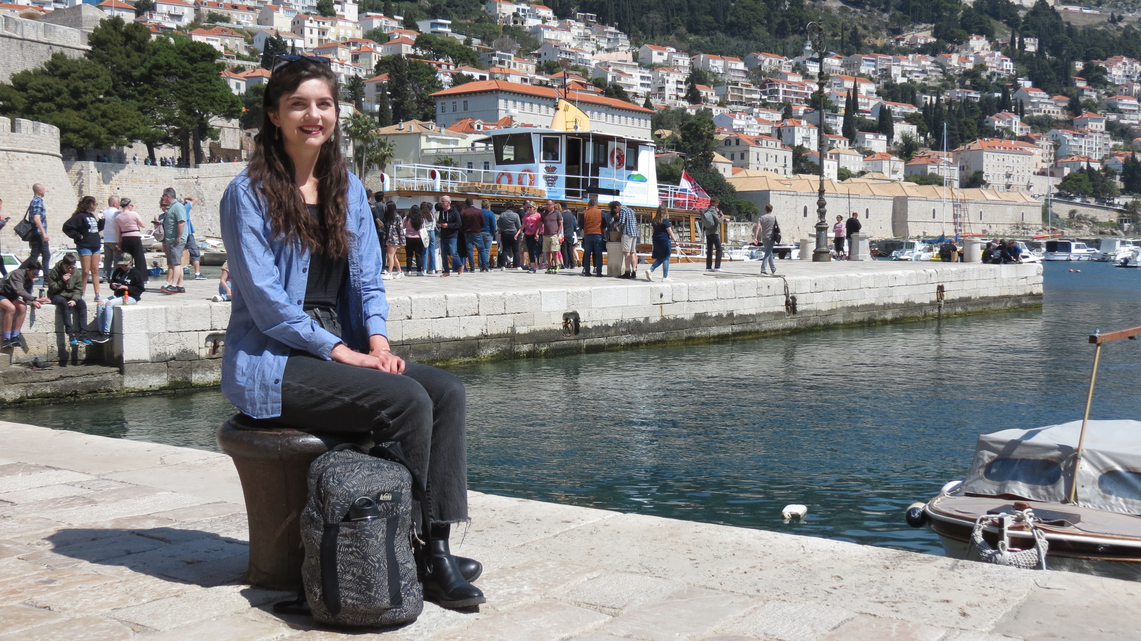 Yvonne Worden sitting on a bollard at Dubrovnik's Old Port, with boats and tourists behind her