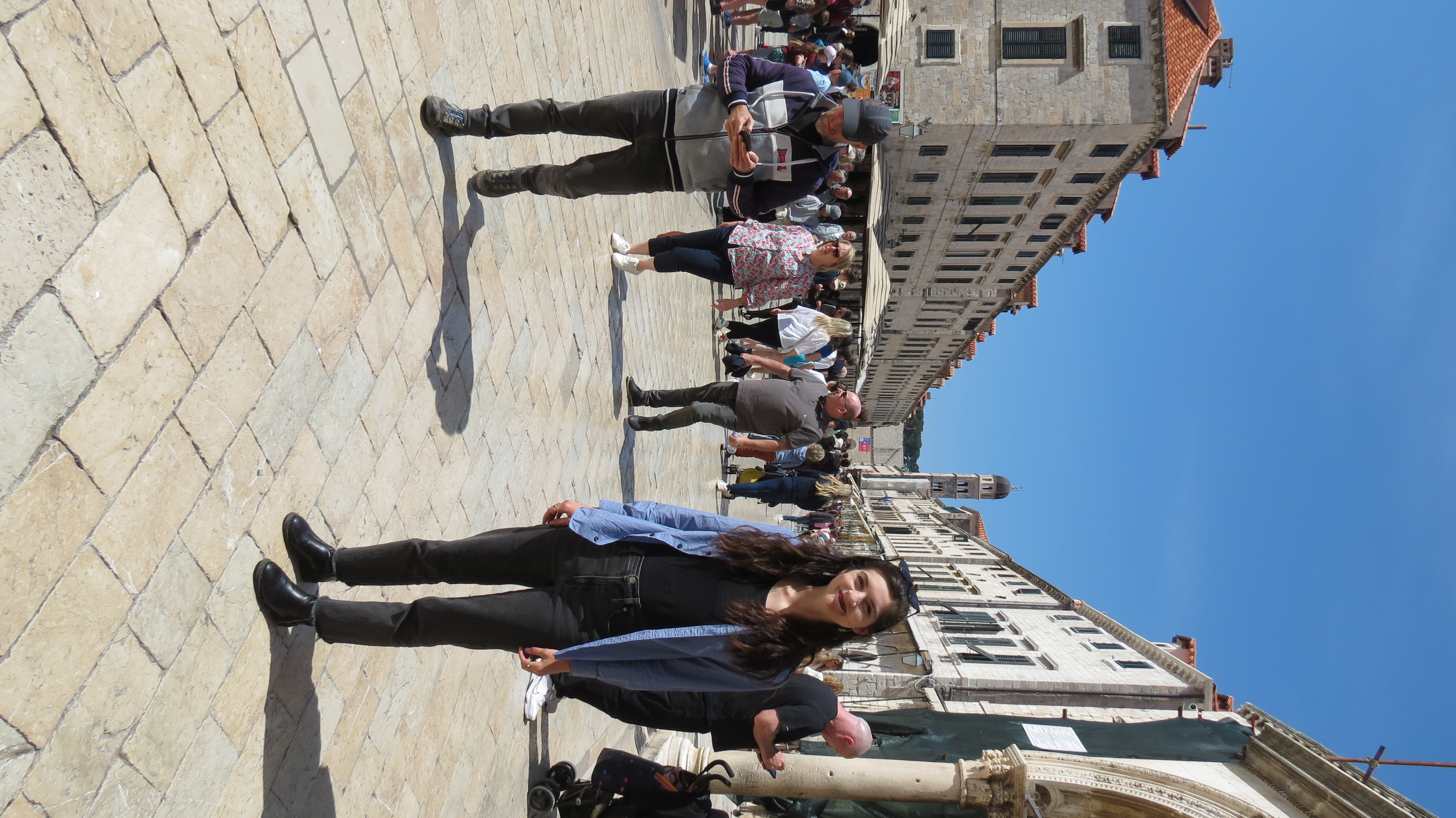 Yvonne Worden standing on Stradun, with a bell tower at the end of the busy street