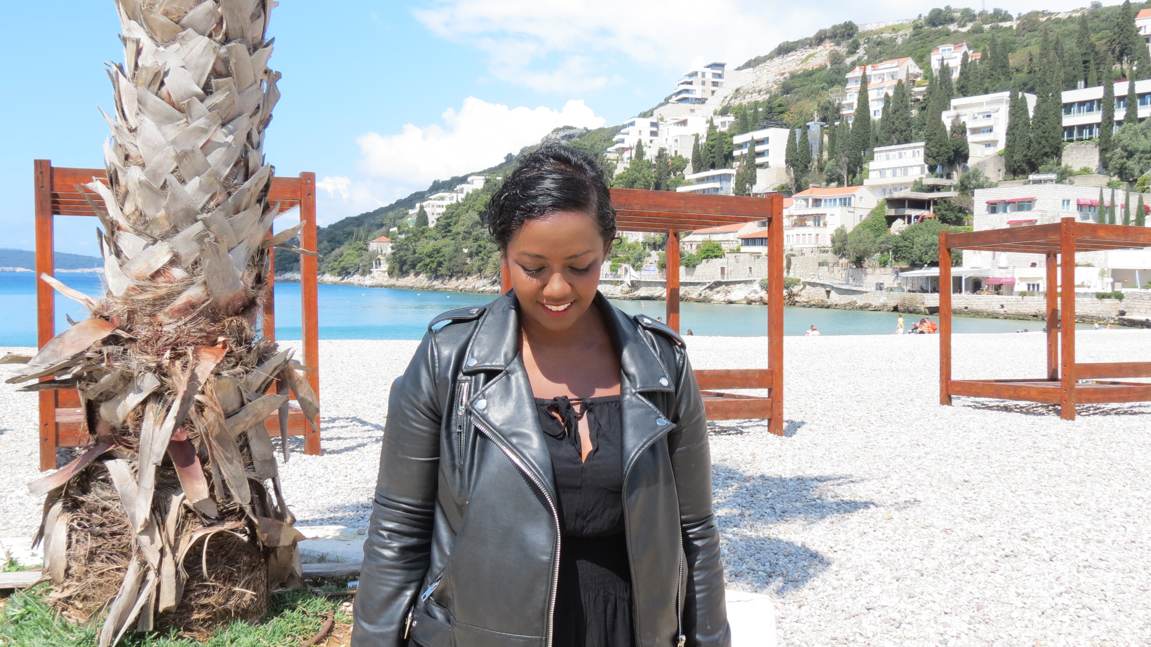 Mita Carriman in a leather jacket smiling on a pebble beach in Dubrovnik, with wooden sunbed frames behind