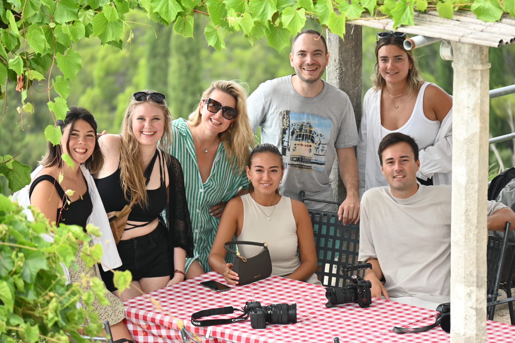Group of smiling digital nomads with cameras at a table under a vine-covered pergola