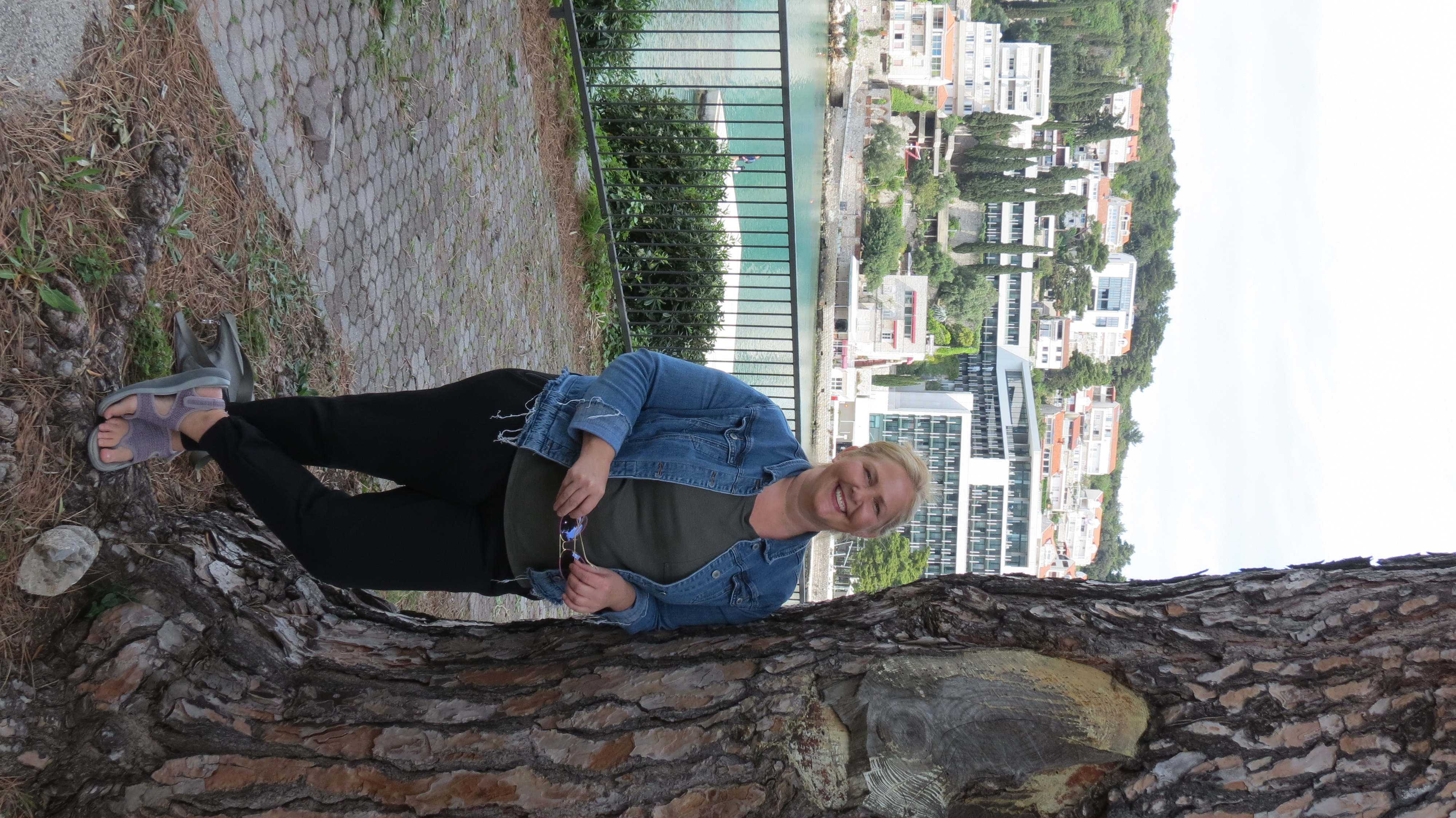 Kristie Sullivan leaning against a pine tree by the sea in Dubrovnik, with hotels on the hillside behind