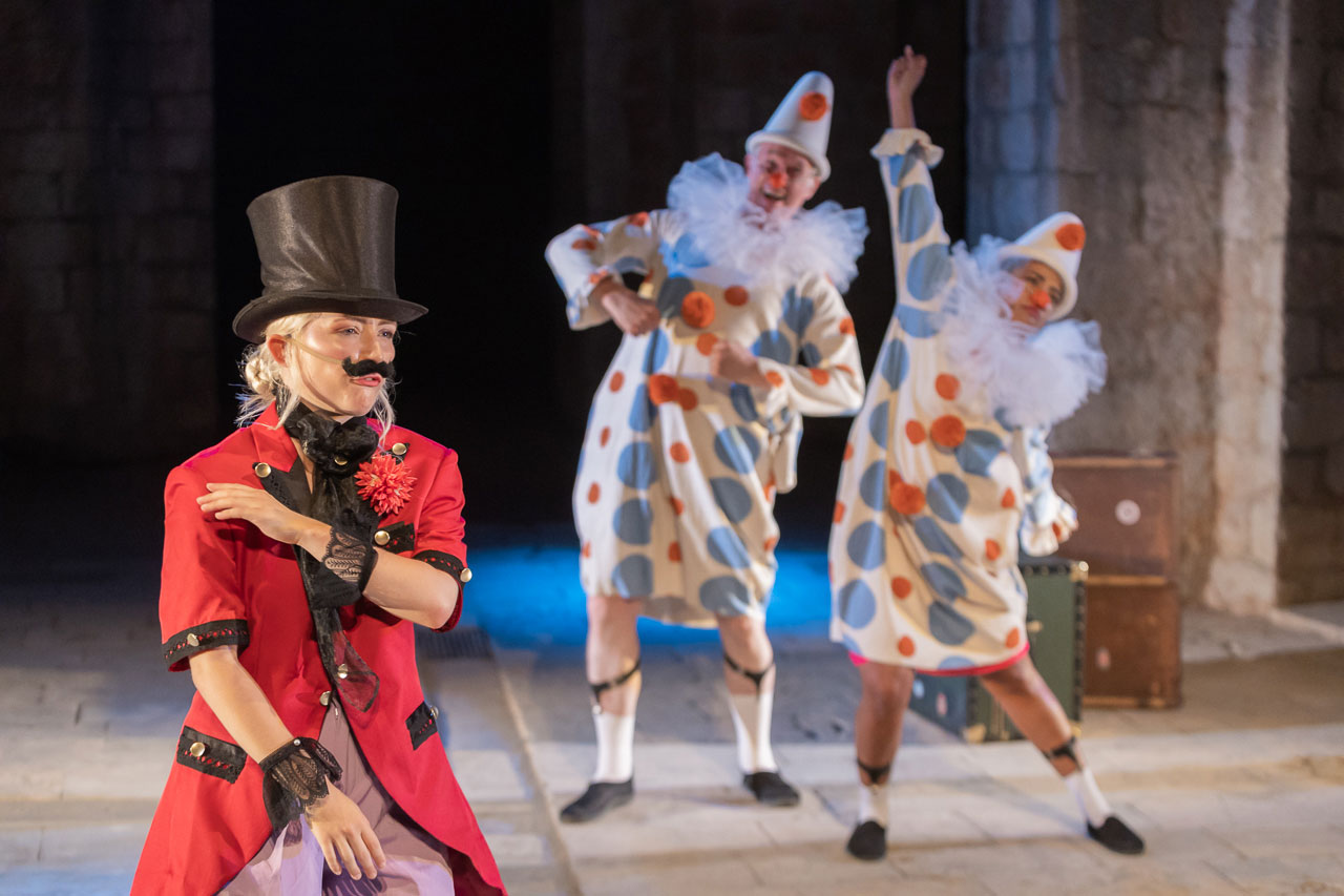 Actress as a ringmaster in a red coat and top hat, with two dancing clowns behind, in Around the World in 80 Days