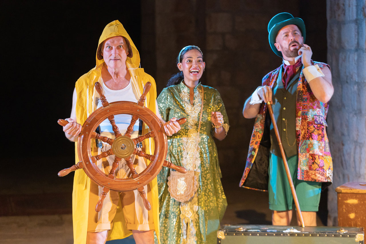 Three actors in colourful costumes, one holding a ship's wheel, in Around the World in 80 Days
