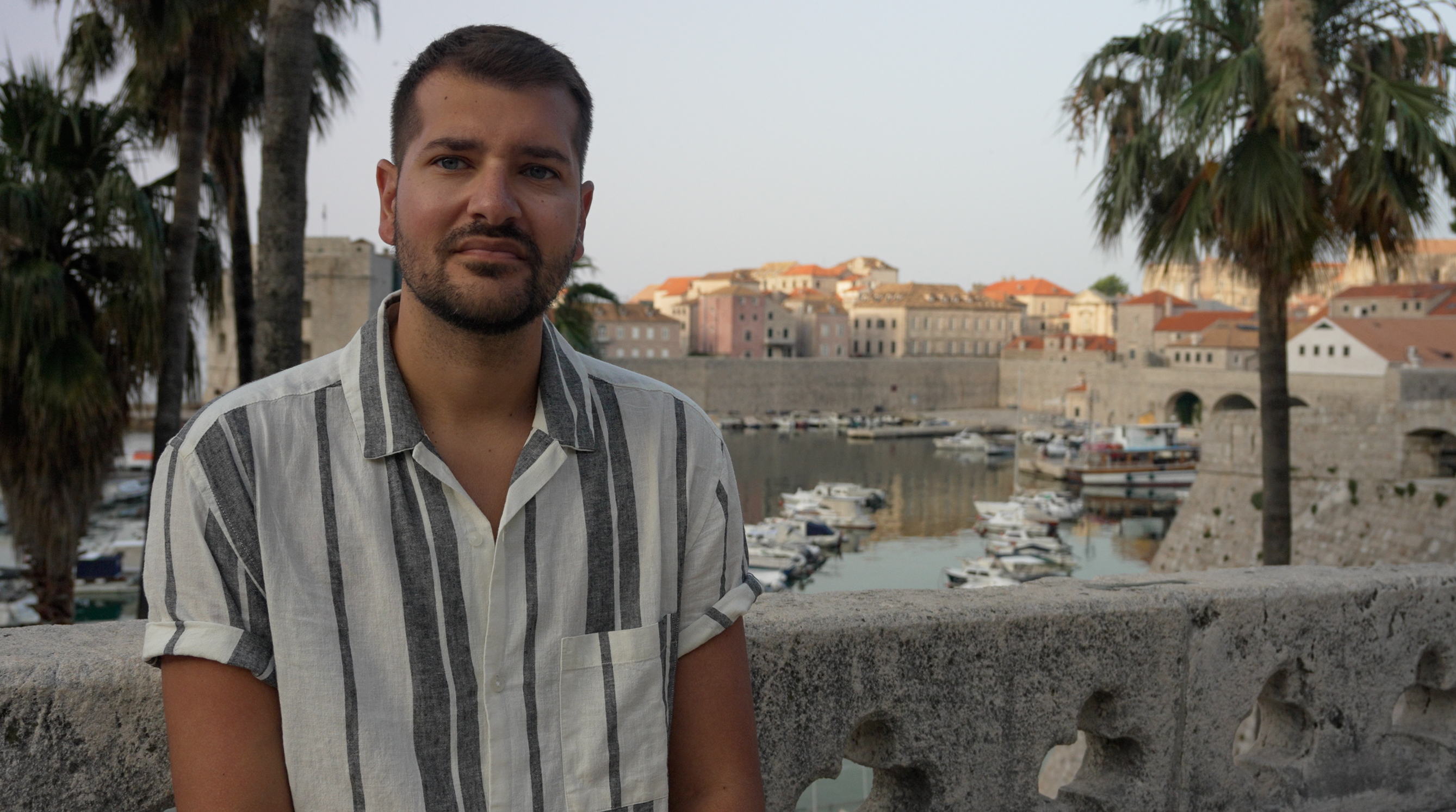 Daniel Hogston by a stone wall overlooking Dubrovnik's Old Port and palm trees at dusk