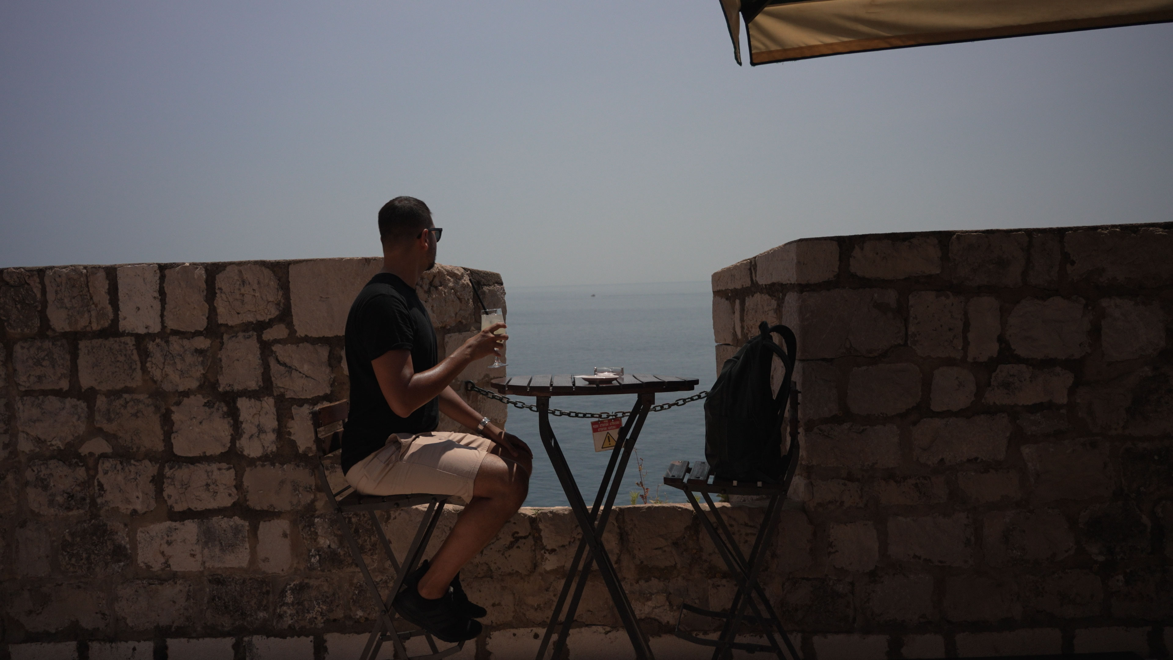Daniel Hogston with a drink at a bar table between stone walls overlooking the sea