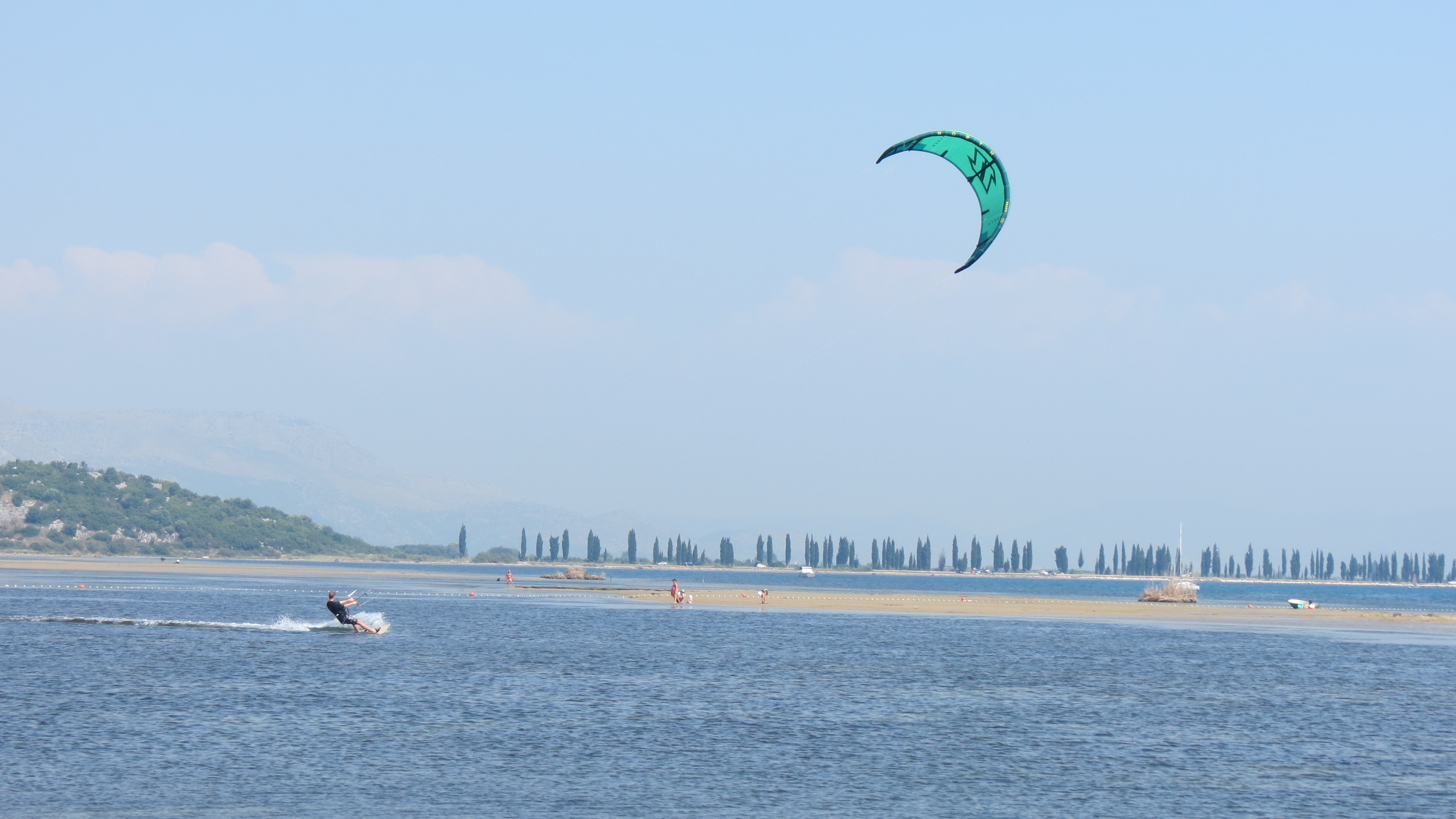Kite surfer with a green kite on shallow water at the Neretva River mouth, sandbanks and cypress trees behind