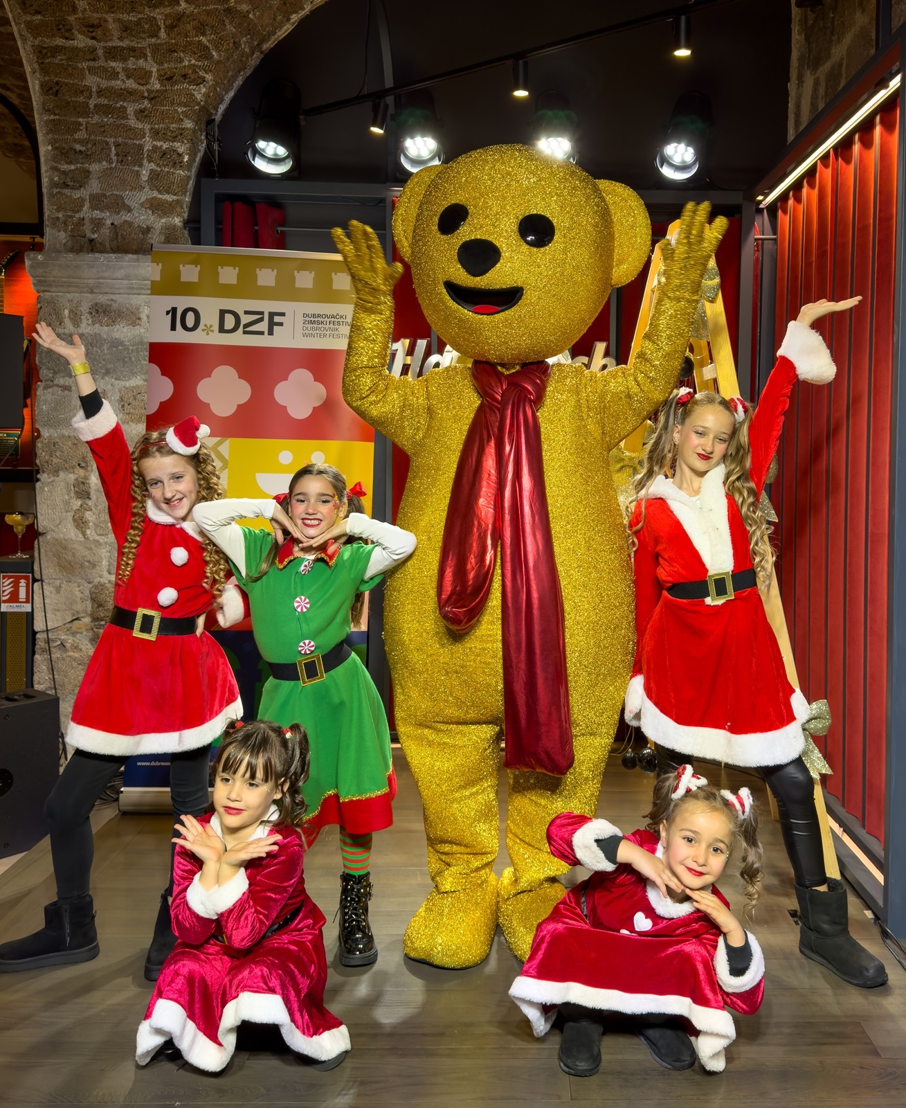 Girls in Santa and elf costumes posing with a golden teddy bear mascot at the Dubrovnik Winter Festival launch