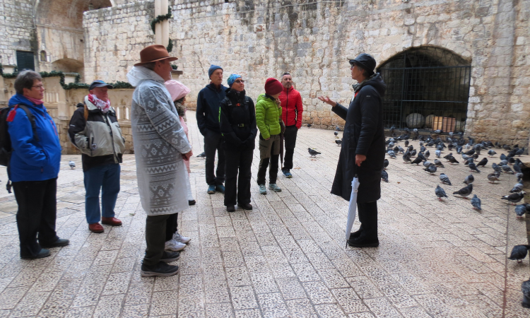 Tour guide speaking to a group in winter jackets in a Dubrovnik square with pigeons
