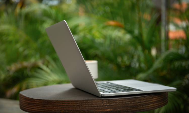 Laptop on a round wooden table with a coffee cup against green tropical plants