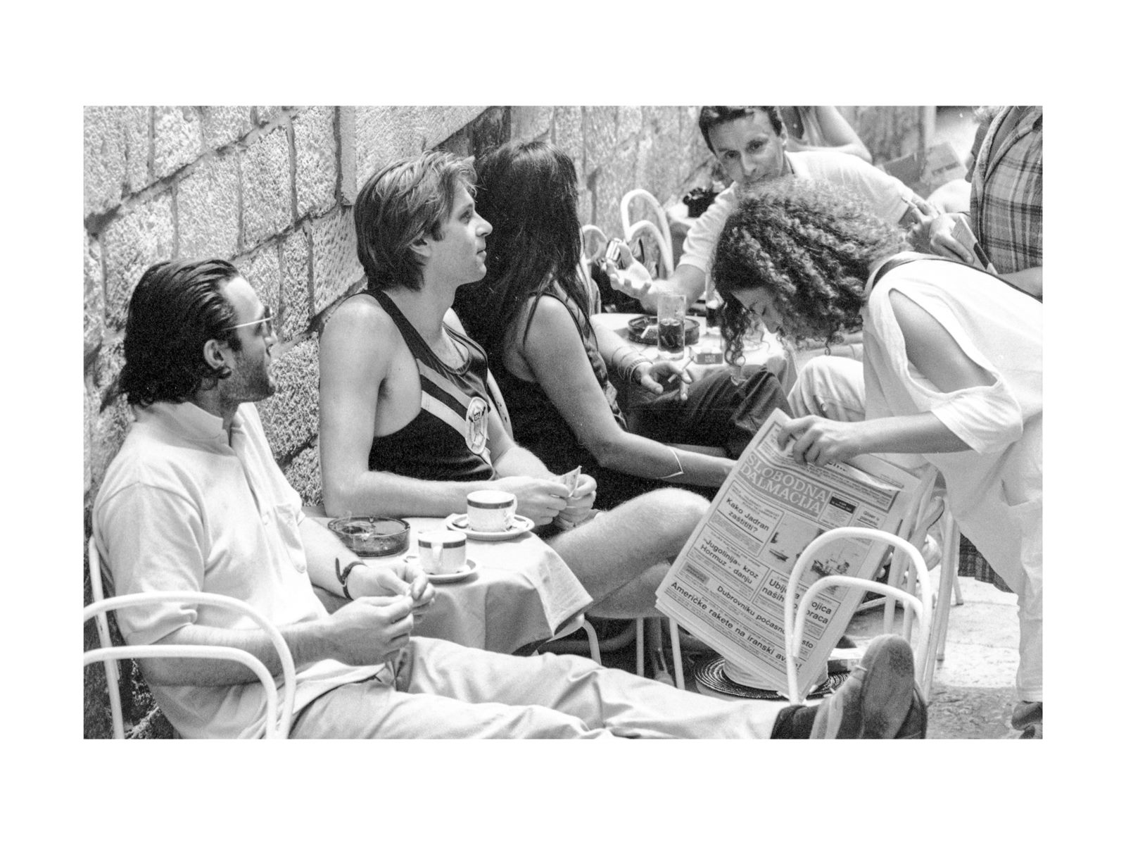 Black-and-white photograph of people at a café table by a stone wall, one woman reading a newspaper