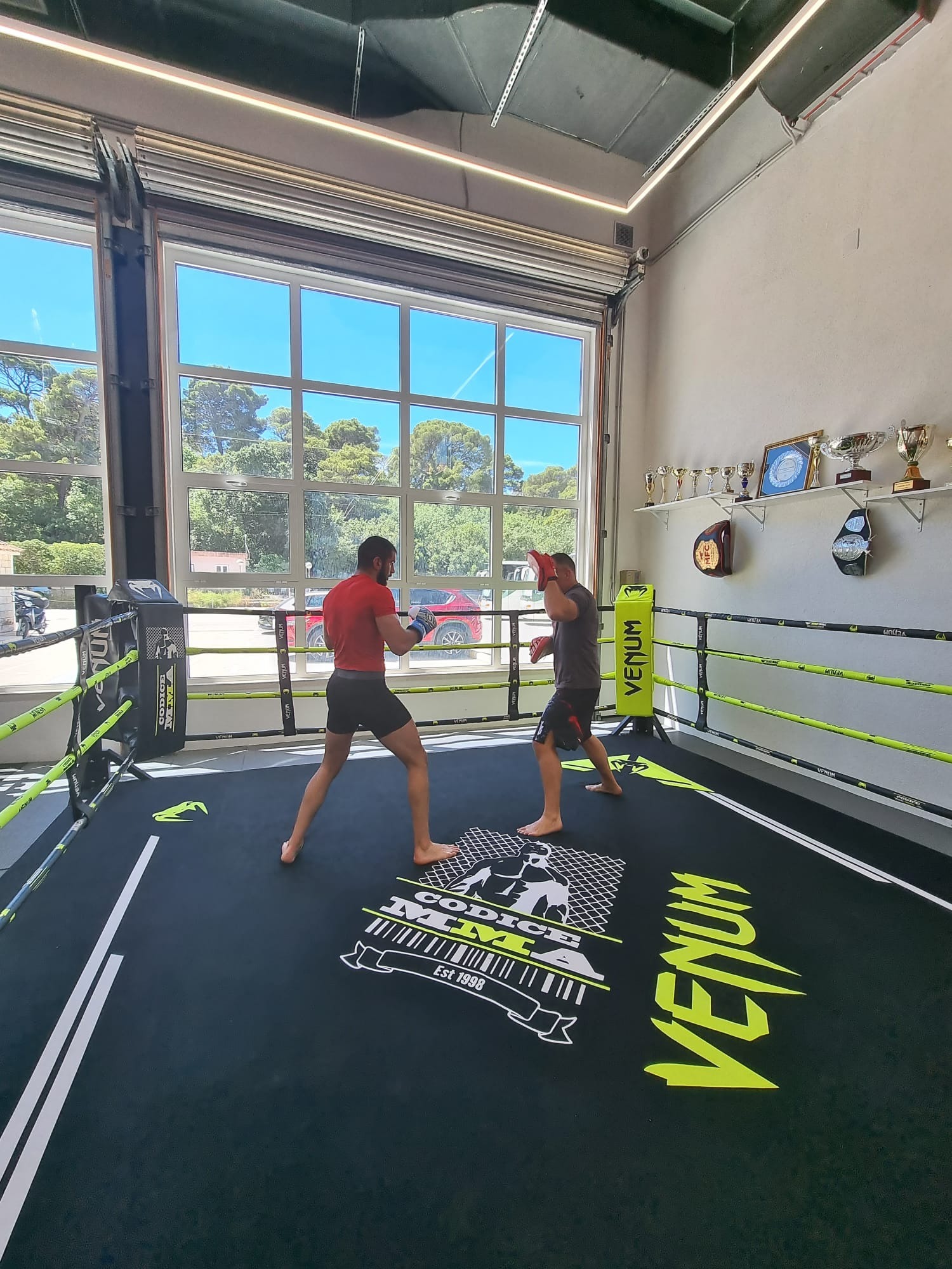 Trainer holding pads for a fighter in a boxing ring, with trophies on the wall