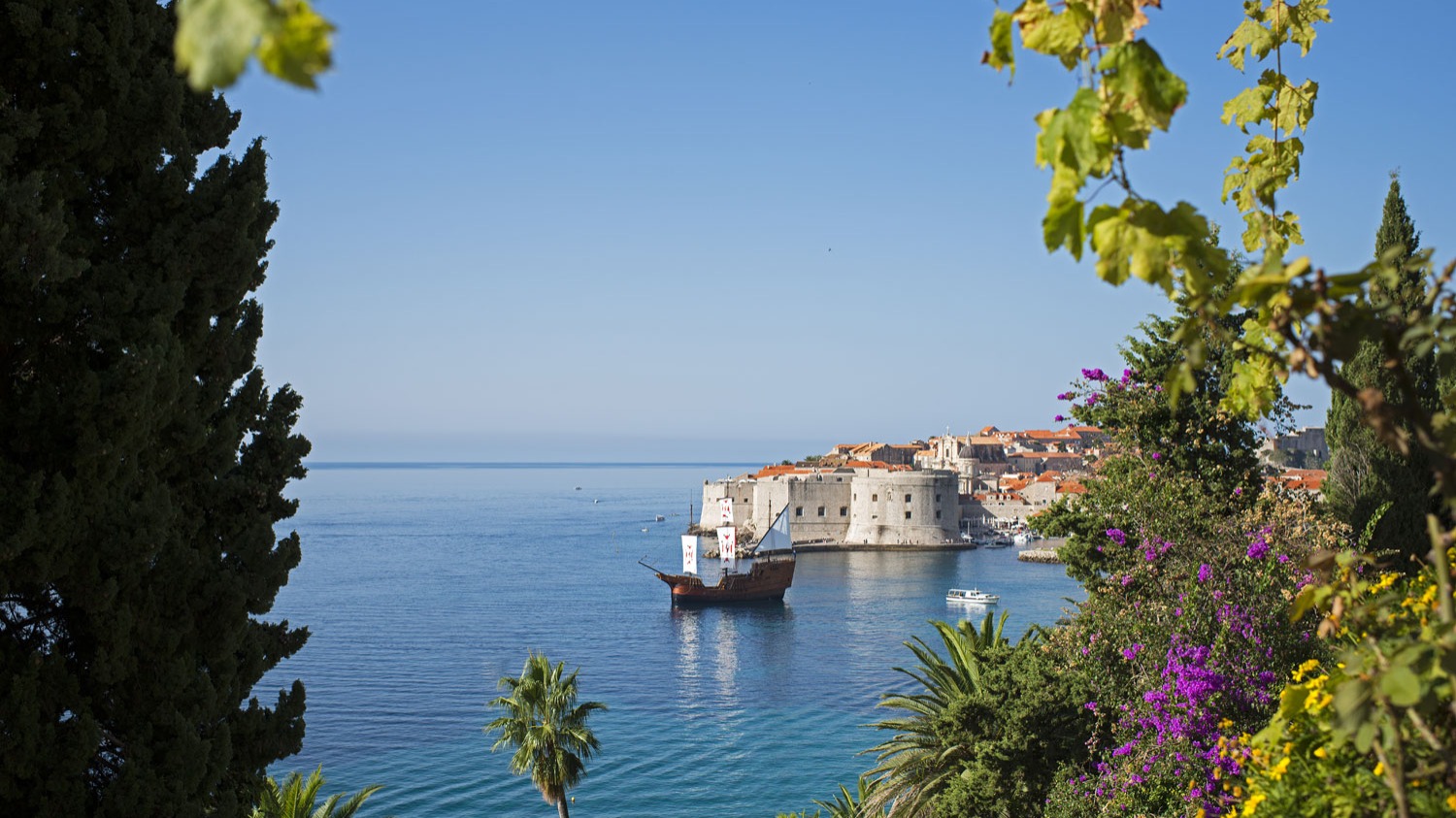 Sailing ship heading toward St John's Fortress and Dubrovnik Old City, framed by trees and flowers
