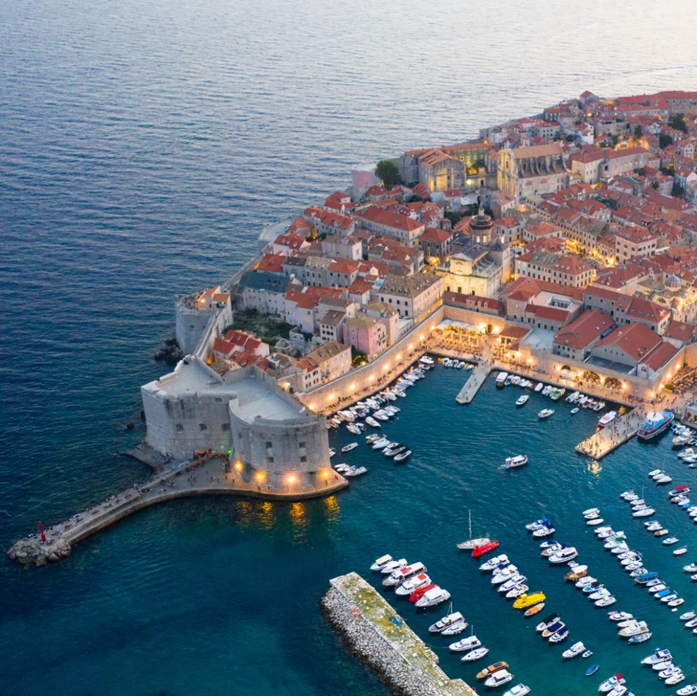 Aerial view of Dubrovnik Old City and its old harbour at dusk, with lit walls and moored boats